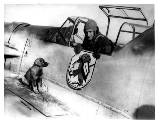 The pilot of a Messerschmitt Bf 109E and his dog mascot admire unit badge used by I Gruppe of JG 51. The legend “Gott Strafe England” translates as “God Punish England. The weeping crow with umbrella pokes fun at British Prime Minister Neville Chamberlain.......